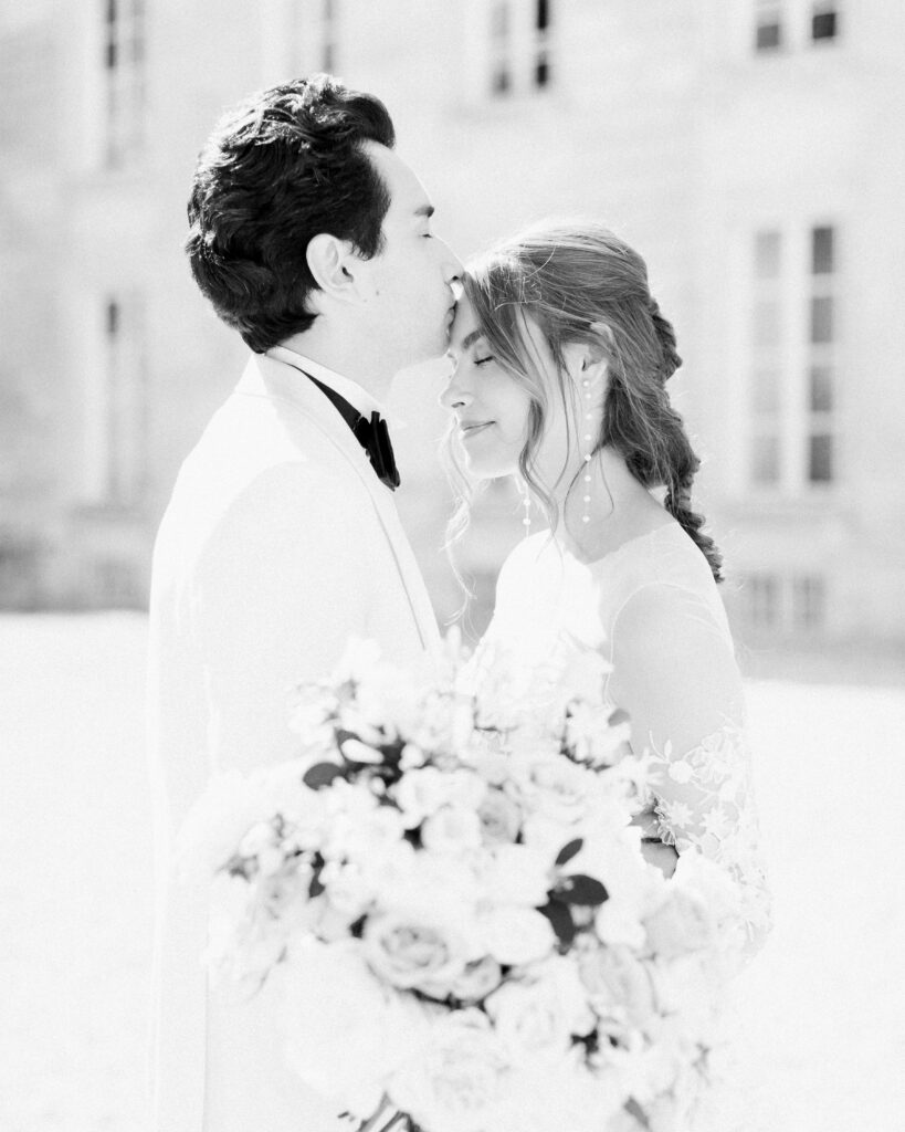 Black and white image of couple embracing, man kissing her gently on the forehead, standing in front of a french chateau wedding venue.