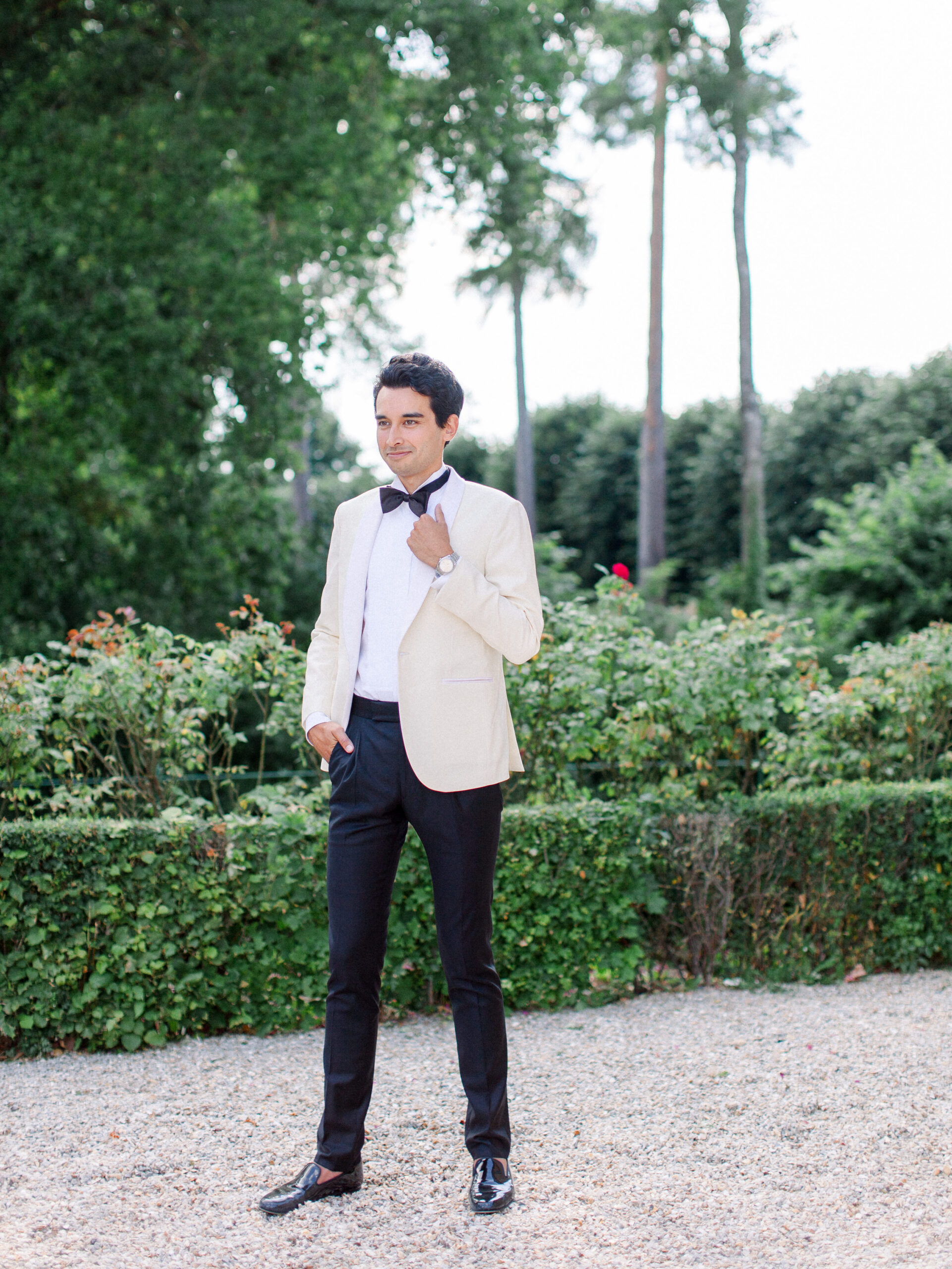 Groom in a white coat and black bow tie, holding lapel in front of rose garden on the grounds of a french chateau wedding venue