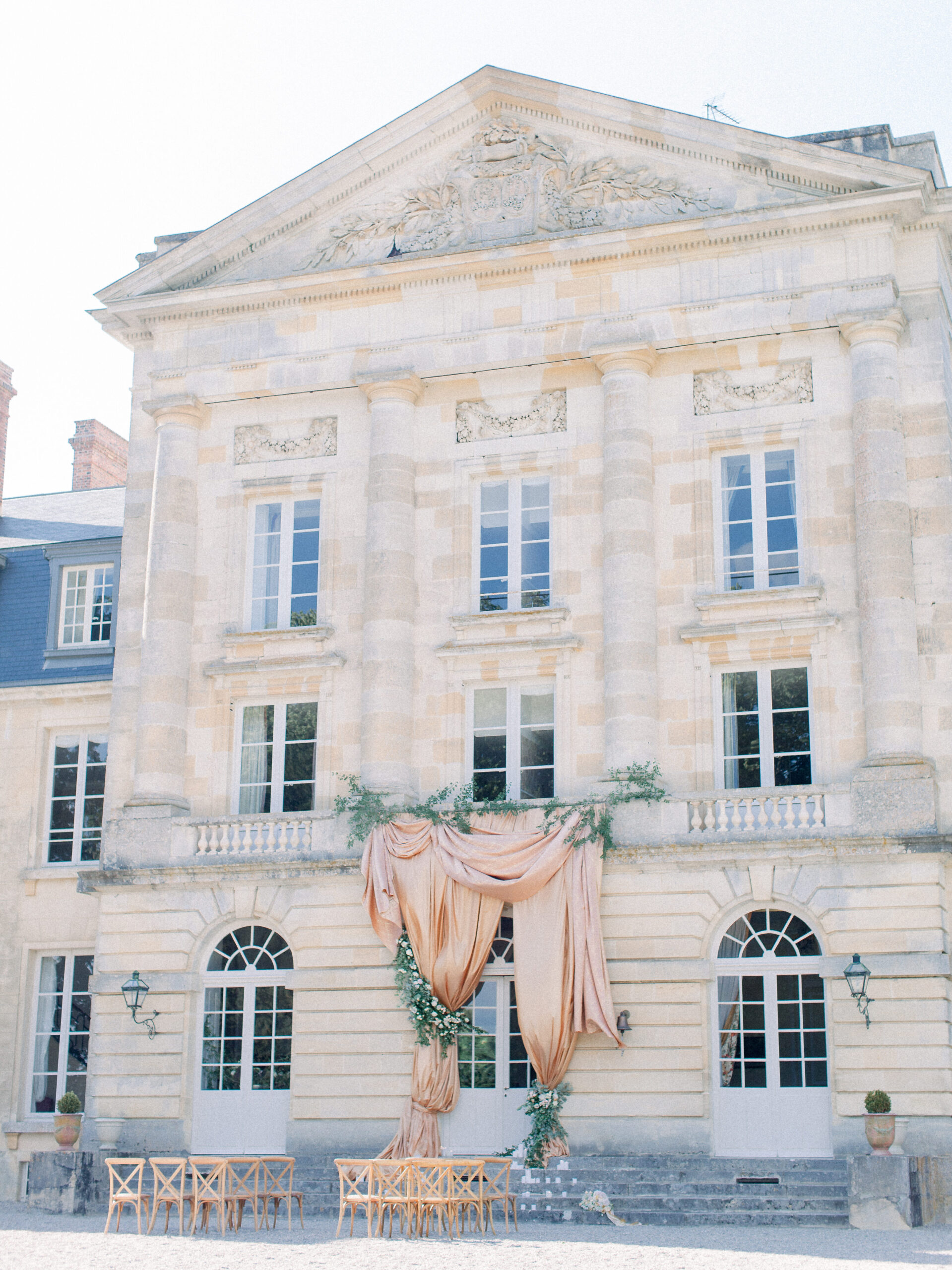 French chateau wedding venue with bistro chairs set for a wedding ceremony and copper silk drapery and lush greenery floral installation. 