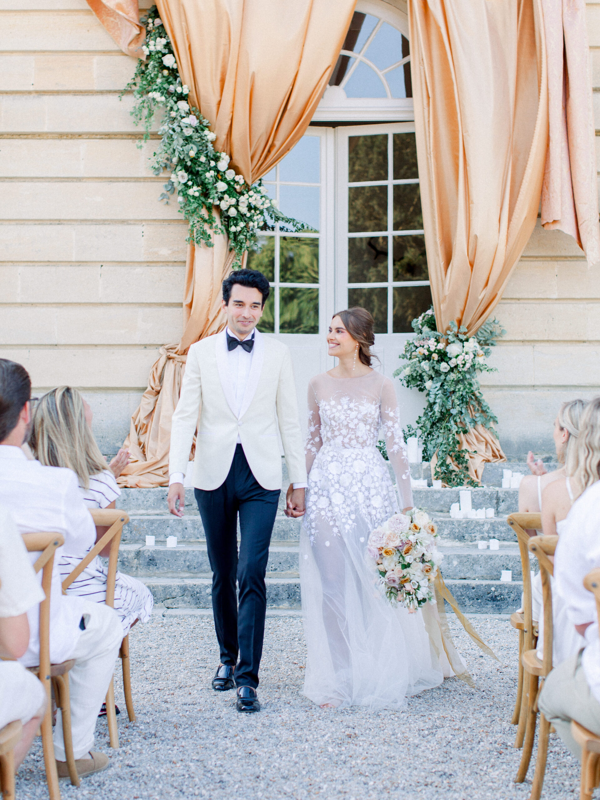 Bride in a long sleeve, translucent wedding gown, and a groom in white coat and black bow tie, walking down the aisle in front of a french chateau wedding venue