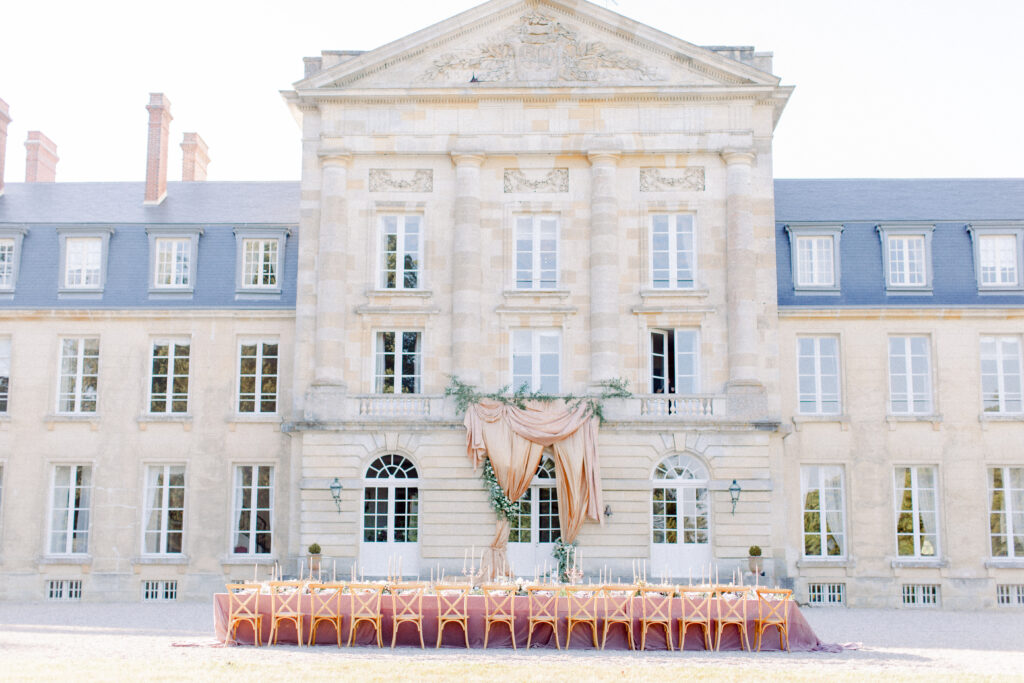 French chateau wedding venue front facade with long head table in front, set with purple velvet table cloth, silver candelabras, and bistro chairs on a summer evening in France.