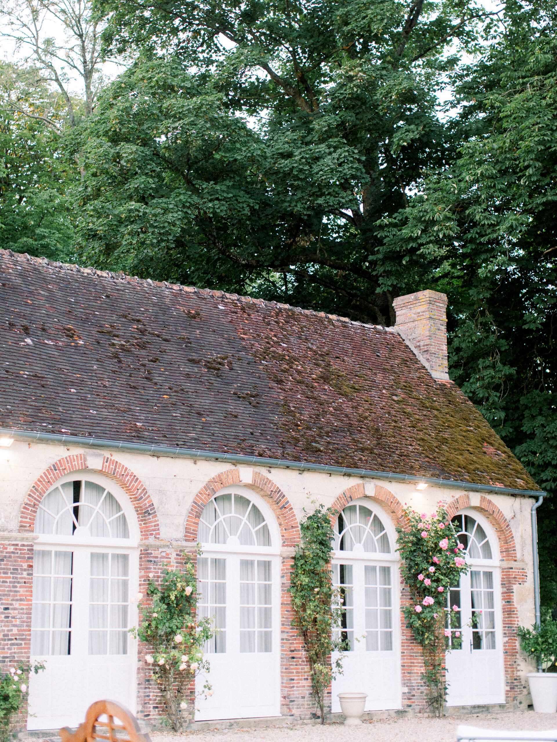 18th century Orangerie with climbing roses, oversized arched windows, and stone roof, on the grounds of a french chateau wedding venue.