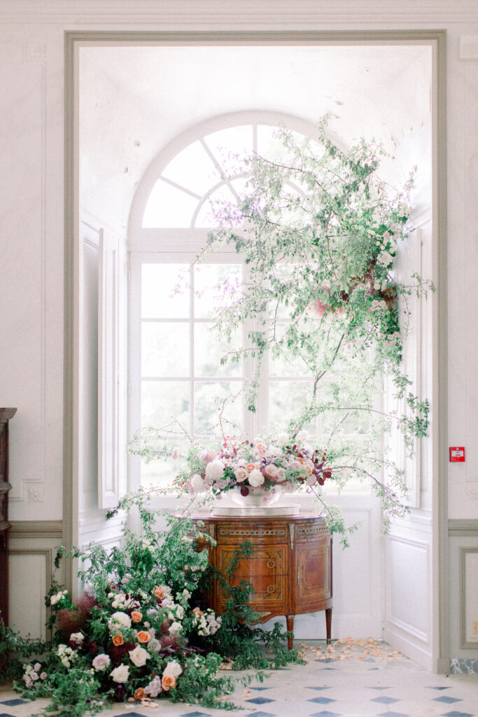 Oversized arched window with antique buffet, stone floors, and floral installation inside a french chateau wedding venue.