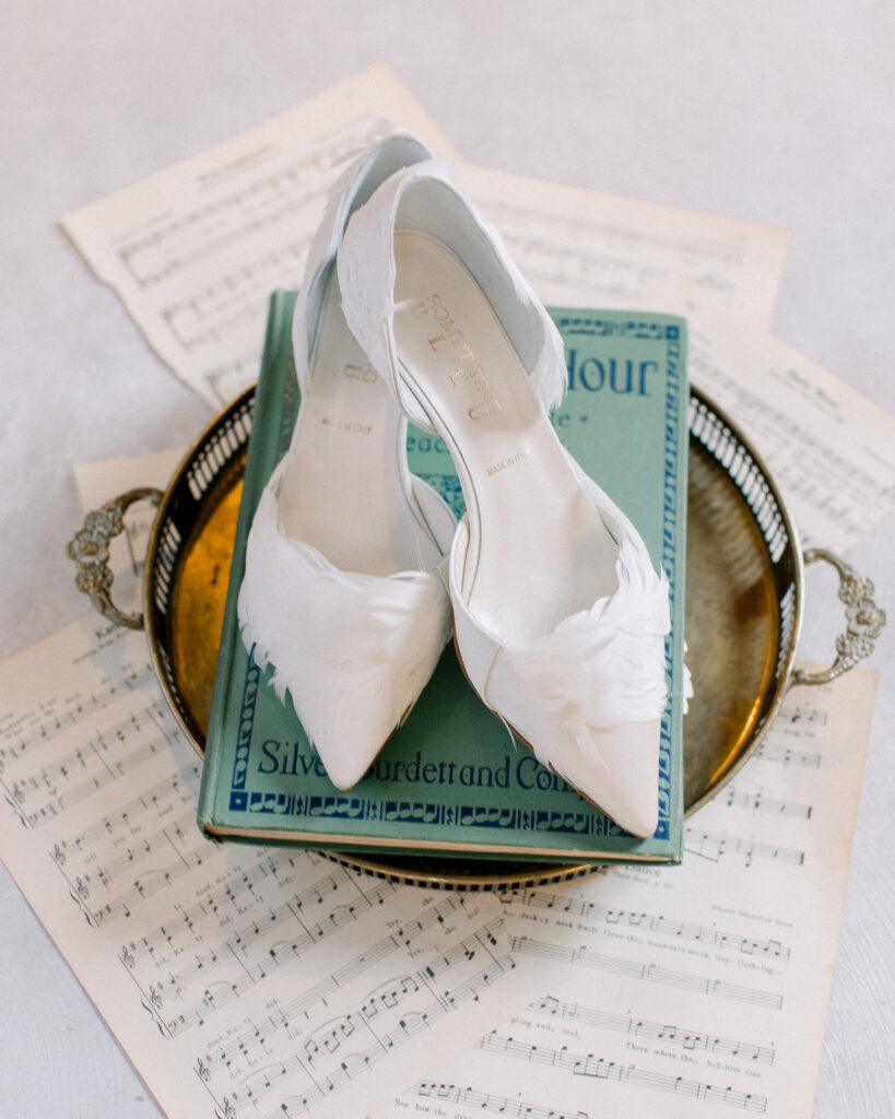 white bridal shoes with feather details, laying on top of a vintage book and silver platter, with sheet music scattered benaeth, wedding day details by Boston wedding photographer Ashley Helen